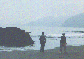 Snapshot of 2 figures by a big boulder on the shore.