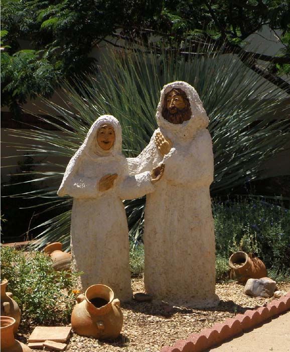 Photo of rough white plaster statues of Mary and Joseph against a background of a large desert plant. There are some potters vessels strewn about.